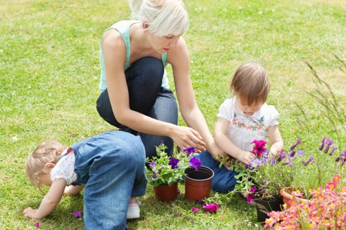 A woman with short blonde hair, wearing a light blue sleeveless top and dark blue jeans, is kneeling on a well-maintained green lawn in a garden, engaging in gardening activities alongside two young children. One child, dressed in a white top and blue dungarees, is bent over, closely examining or picking flowers close to the ground, while the other, also in a white top with floral patterns, is seated nearby, tending to a flower bed with purple, pink, and orange blooms. The garden features a lush, evenly cut grass area with a border of vibrant flowering plants and potted plants, including small containers and larger pots. In the background, there are trees and shrubs providing a natural environment, suggesting a quiet and private outdoor space likely part of a residential garden. The scene appears to be taking place during daytime with soft, natural light, indicative of good weather. This image highlights activities such as planting, flower tending, and outdoor family gardening, aligning with professional garden care and landscaping services available from Hedge Trimming Mottingham in the Mottingham area, within the London borough or nearby postcode district.
